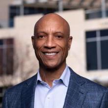 A person in a suit smiling in front of a modern building.