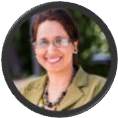 Minny Walia smiling wearing glasses and an olive green blazer