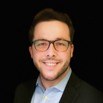 A person wearing glasses, a suit, and smiling against a black background.