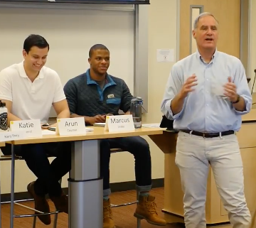 Three individuals sitting and one standing in a conference room setting.