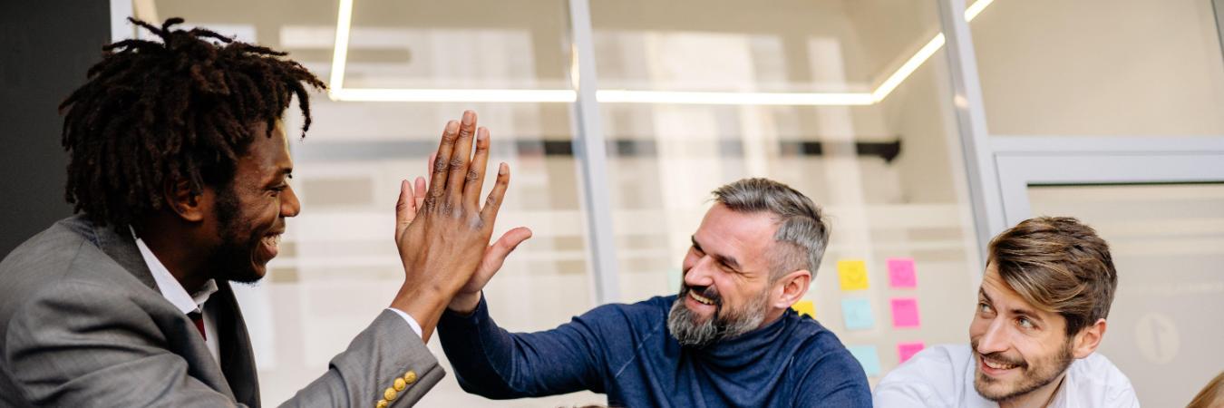 Five coworkers smile and gather around a laptop in an office as two men high-five.