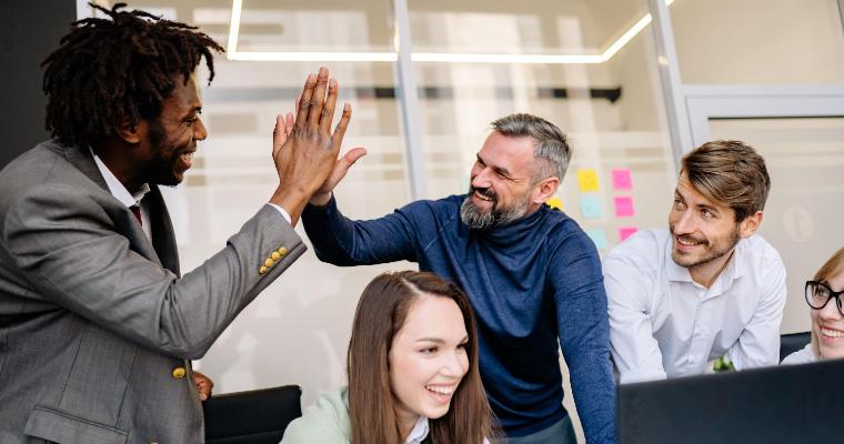 Five coworkers smile and gather around a laptop in an office as two men high-five.