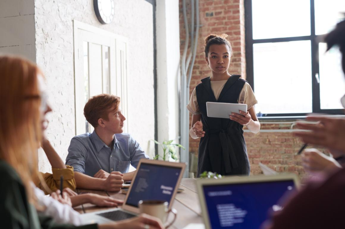 Coworkers meet around a conference table while a woman standing at the center holds a tablet and addresses the group.