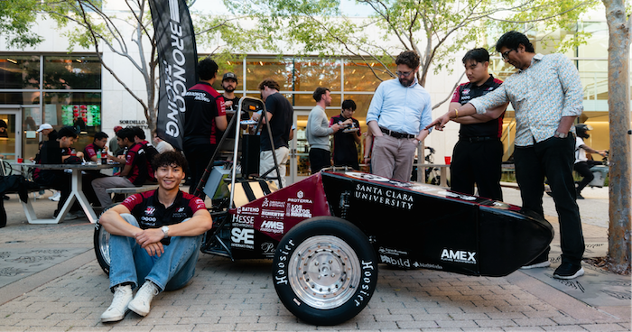 A Santa Clara University open-wheel race car surrounded by team members, with Aiden Abarca seated beside the front wheel and a Bronco Racing banner behind him.