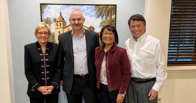 Four individuals pose together in a well-lit room, with a painting of a building and palm trees in the background.