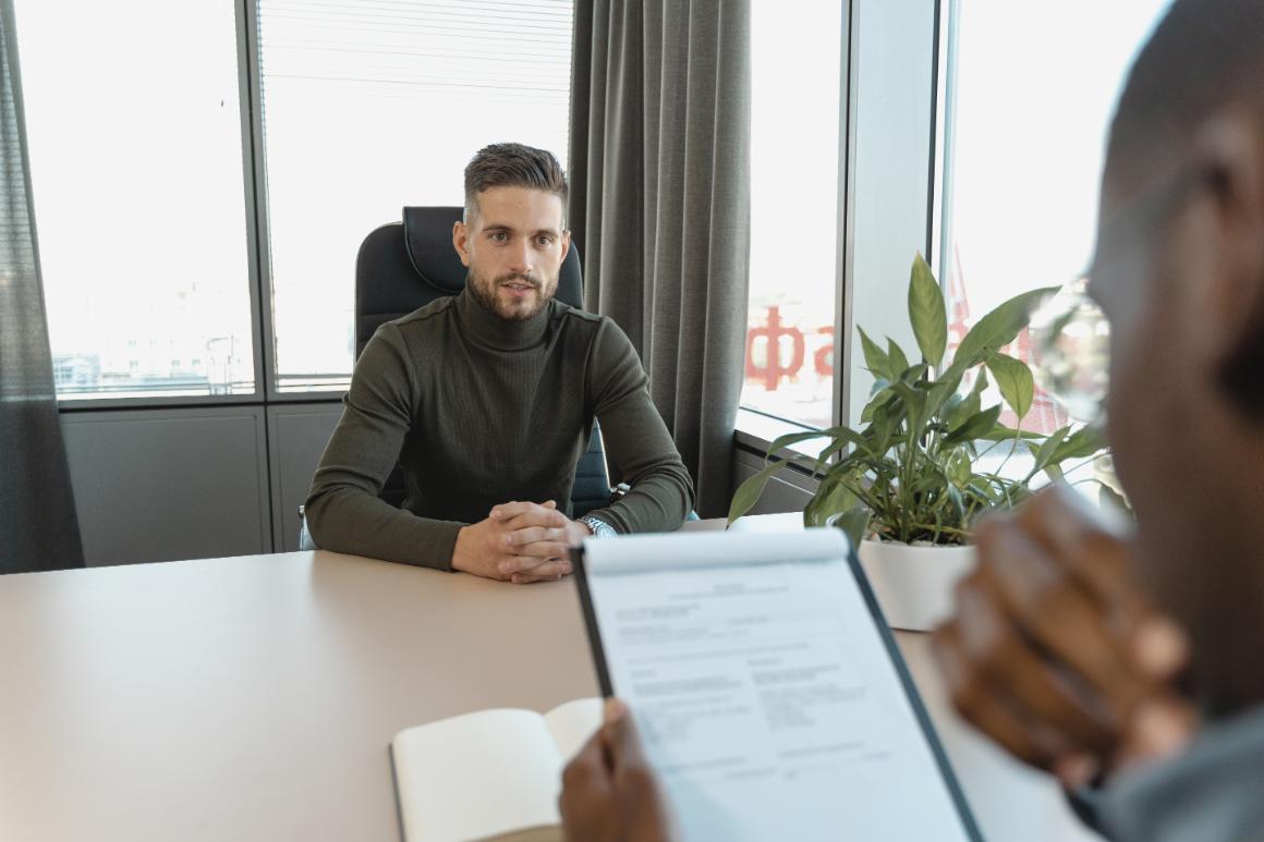 A man in a dark green turtleneck sits at a desk during an interview, with a window and a plant in the background. Another persons hand is holding a clipboard in the foreground.