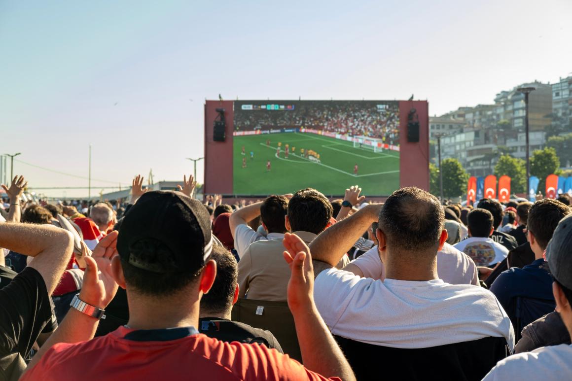 A crowd of fans with raised arms gathers outdoors to watch a soccer game on a large screen, with urban buildings in the background.