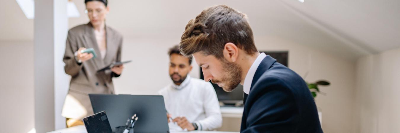 Three people work in a modern office; one man in a suit writes at a desk, another man uses a laptop, and a woman stands in the background holding a tablet.