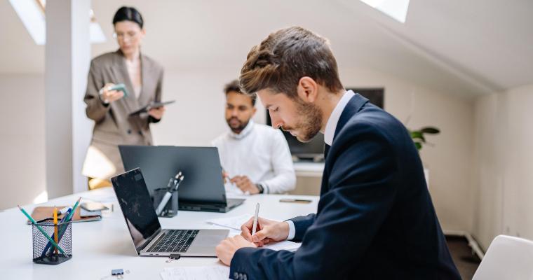 Three people work in a modern office; one man in a suit writes at a desk, another man uses a laptop, and a woman stands in the background holding a tablet.