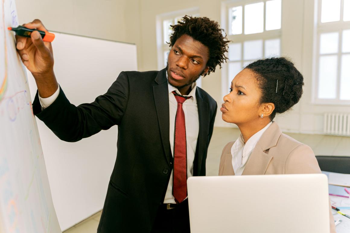 Two people in business attire stand by a whiteboard; one writes with a marker while the other looks on thoughtfully, with a laptop in the foreground.