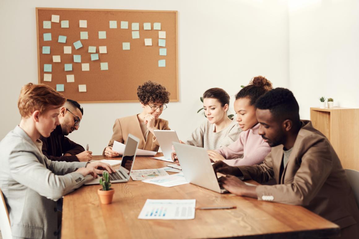 Six adults collaborate around a conference table with laptops, papers, and tablets in a bright office.