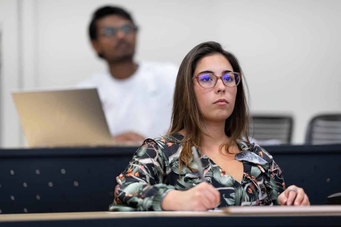 A young woman with long brown hair and glasses seated in a classroom, focused on her notes. A male student is partially visible in the background, working on a laptop.