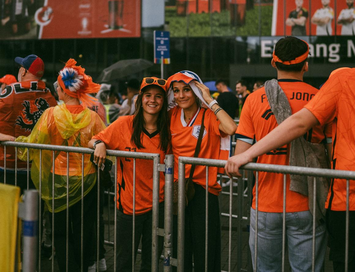 Two young women in orange shirts pose with a metal fence in a crowded outdoor area, smiling for the camera. The scene is lively, with many others around them also wearing orange.