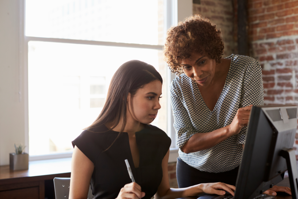 Two women collaborate at a computer in a sunlit office, with one pointing toward the monitor.