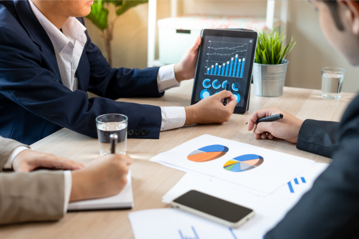 Three professionals in business attire are gathered around a table, reviewing data on a tablet and papers with charts during a meeting.