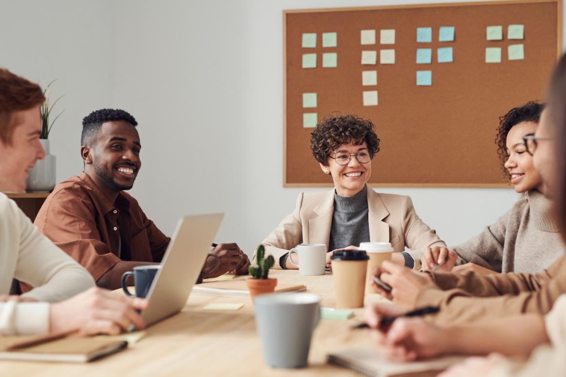 Five people gathered around a table with a laptop, notebooks, coffee cups, and a corkboard covered in sticky notes.