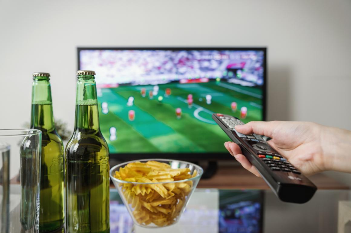A hand holds a remote control in front of a TV displaying a soccer match, with a bowl of chips and two green bottles on a table.