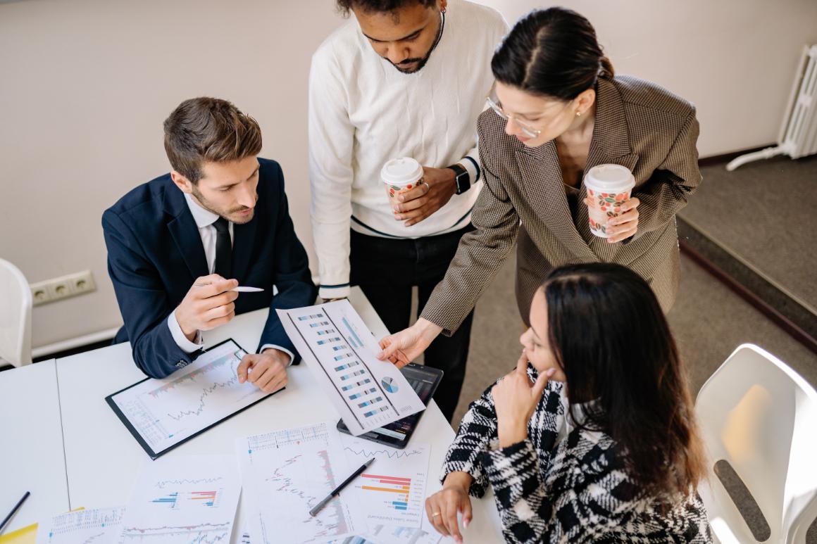 Five colleagues review charts and data around a table, with printed reports, laptops, tablets, and coffee cups.
