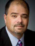 Alt text: Man in a suit and tie with a neutral expression.