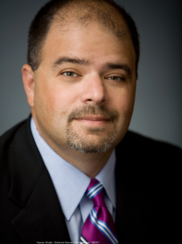 Alt text: Man in a suit and tie with a neutral expression.