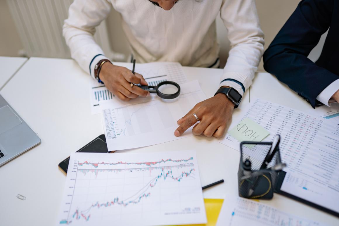A person examining financial reports on a white table using a magnifying glass, with documents, a laptop, and a sticky note that says HOLD visible in the scene.