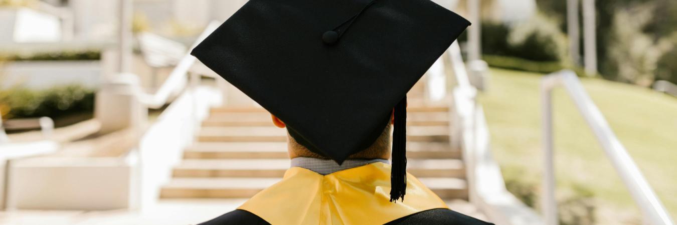 A graduate in a cap and gown with a yellow stole stands facing a sunlit staircase, symbolizing new beginnings and achievement