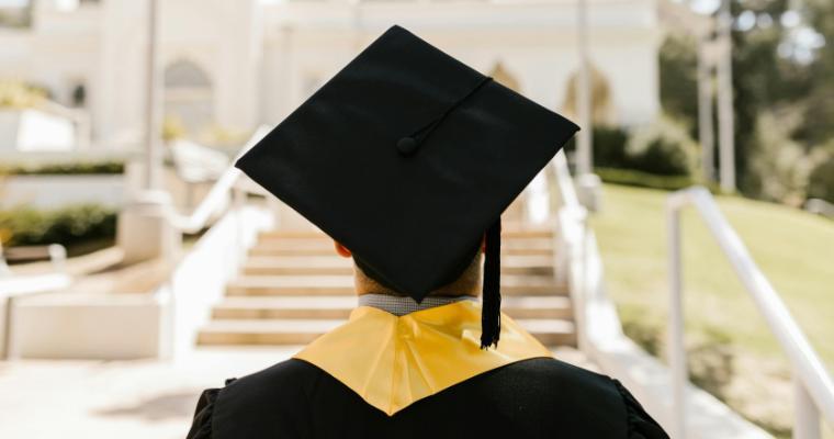 A graduate in a cap and gown with a yellow stole stands facing a sunlit staircase, symbolizing new beginnings and achievement