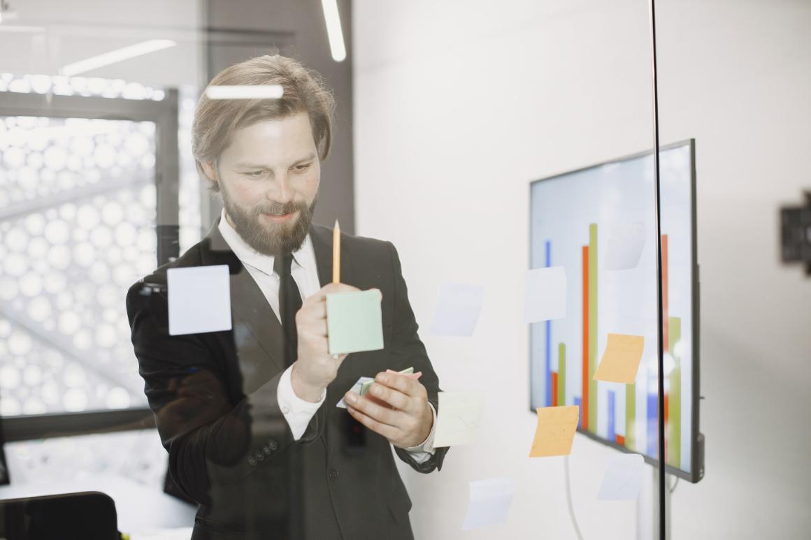 A bearded businessman in a suit stands in an office, placing a light green sticky note on a glass partition in front of a digital bar chart display.
