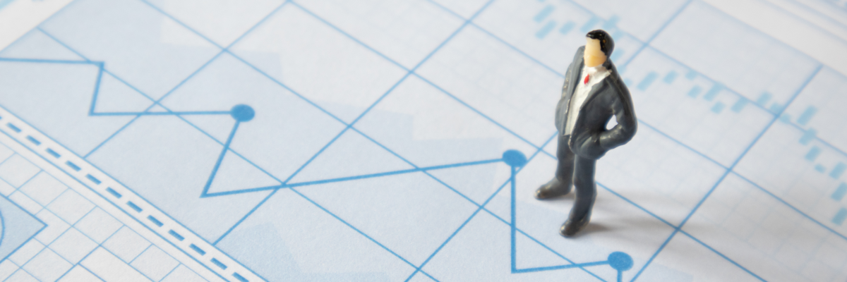 A miniature man in a gray suit stands on a light blue financial chart with various data lines and bars.