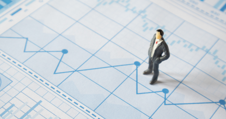 A miniature man in a gray suit stands on a light blue financial chart with various data lines and bars.