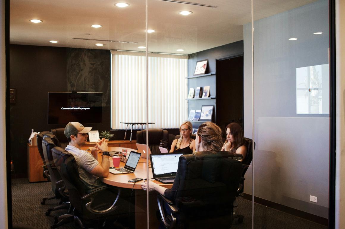 A group of four people in a conference room meeting, with laptops on a circular table and a screen displaying Commercial work in progress.
