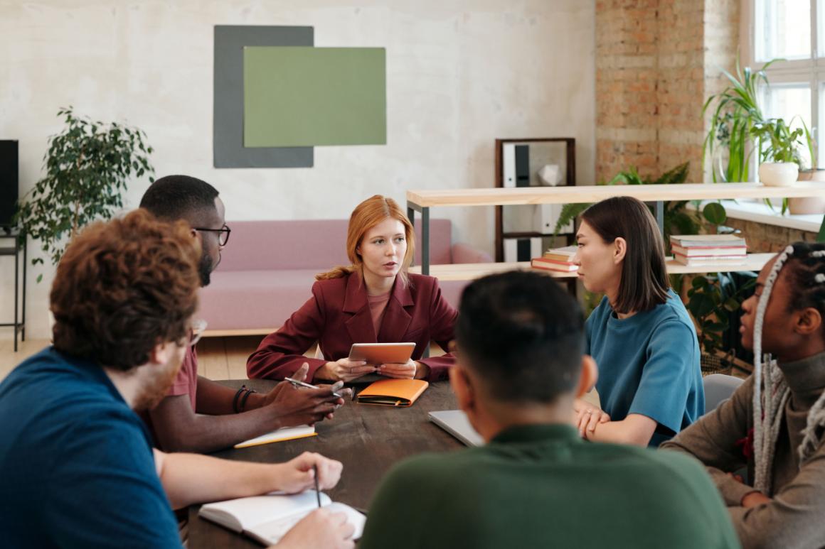 Group discussion around a table in a bright office, with a red-haired woman holding a tablet at the center and other participants taking notes.