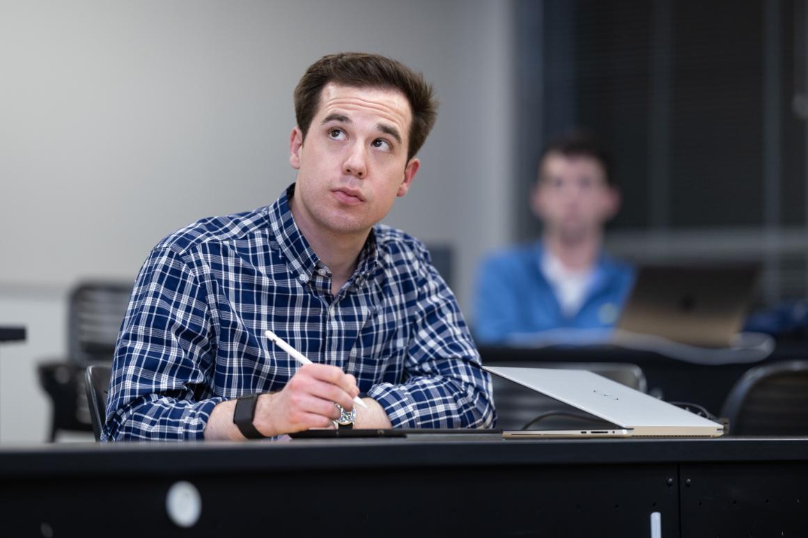 A student in a blue plaid shirt attentively listens and takes notes with a pen in a classroom setting