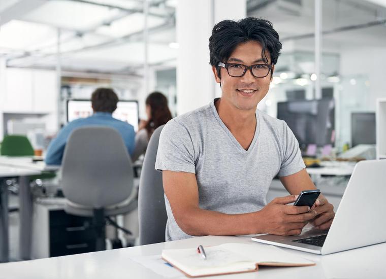Man sitting at his laptop smiling at the camera in an office setting.	