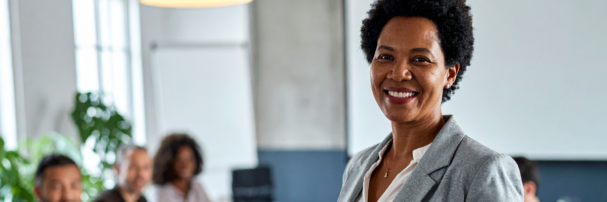 Smiling woman in a gray blazer standing in a modern conference room with colleagues seated at a table behind her.