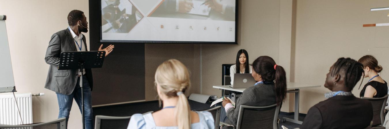 A man presents a business analysis in a conference room, gesturing towards a screen as seated attendees listen attentively.