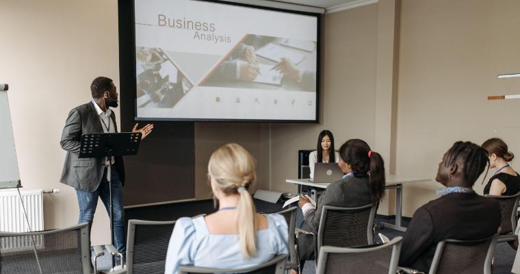 A man presents a business analysis in a conference room, gesturing towards a screen as seated attendees listen attentively.