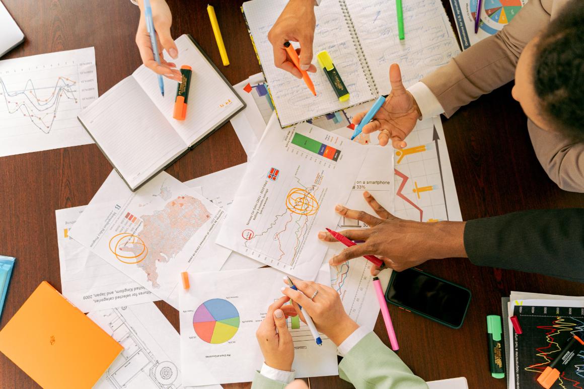 People collaborate over printed charts, maps, notebooks, and colorful markers on a table.