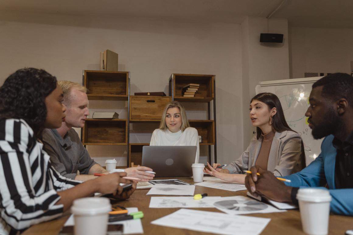 Five adults collaborate around a table with papers, coffee cups, and a laptop in front of shelves and a whiteboard.