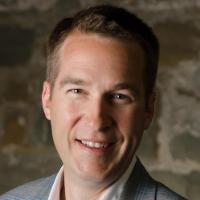 A man in a light gray jacket smiling at the camera.