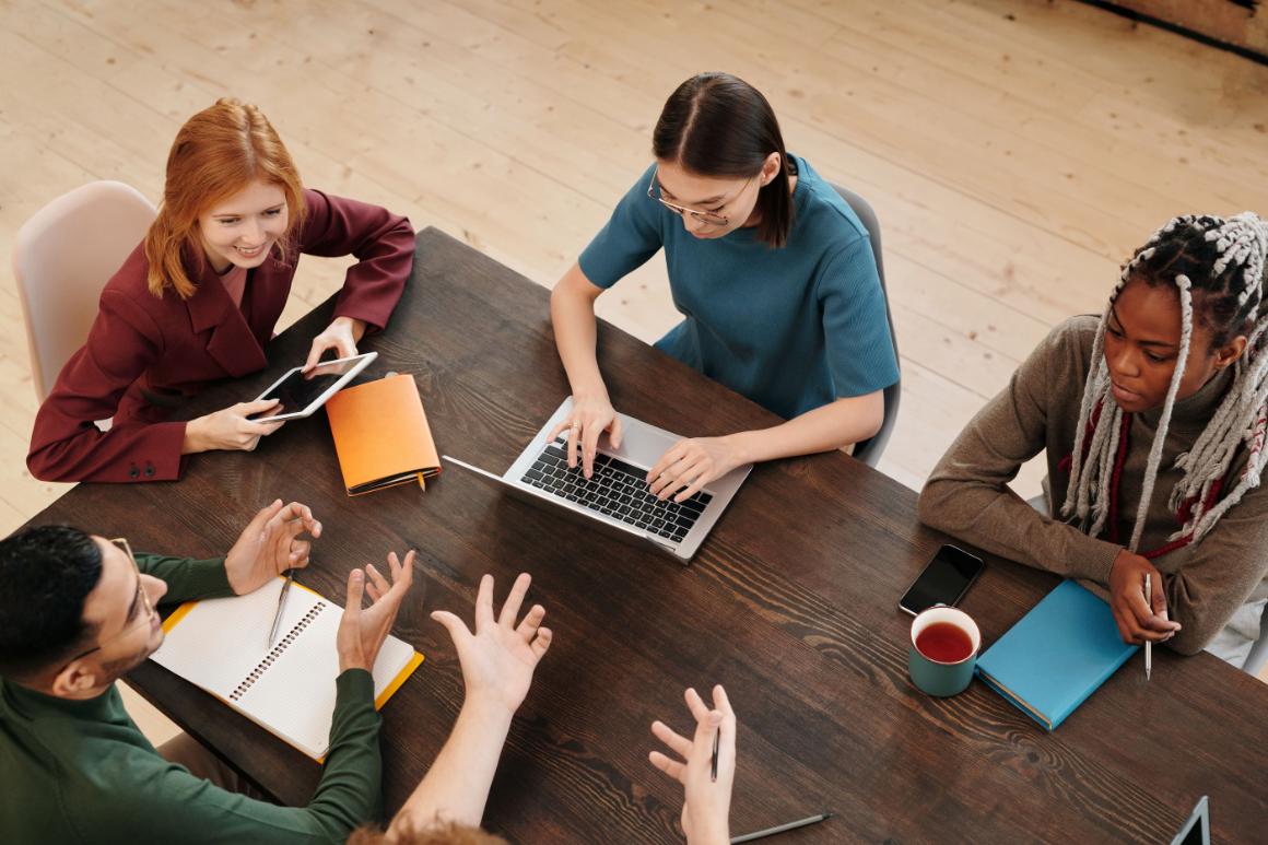 Diverse group collaborating around a wooden table with a laptop, tablet, notebooks, and tea.