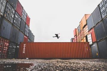 Person jumping between stacks of shipping containers in an industrial area.