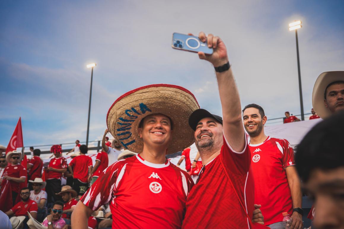 Two men in red sports jerseys joyfully taking a selfie in a stadium filled with cheering fans.