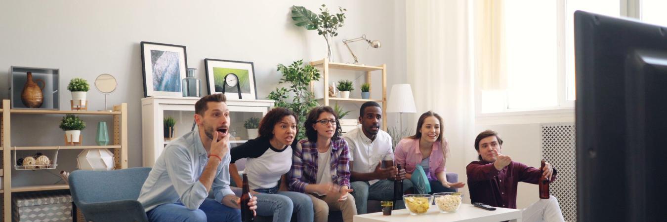 A diverse group of six friends excitedly watching TV together in a cozy living room, with a bowl of popcorn and drinks on the coffee table.