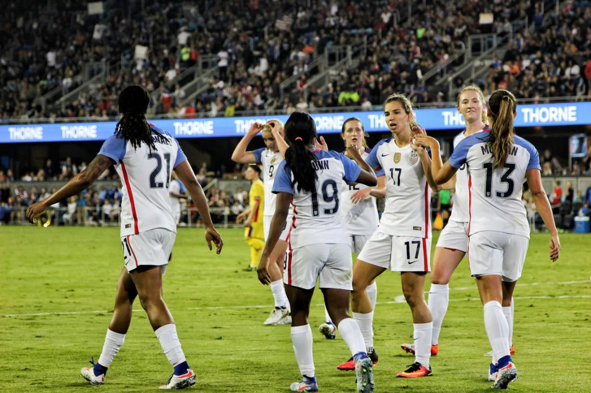 U.S. womens soccer players celebrate on the field, wearing white jerseys with blue and red accents, with a cheering crowd in the background.