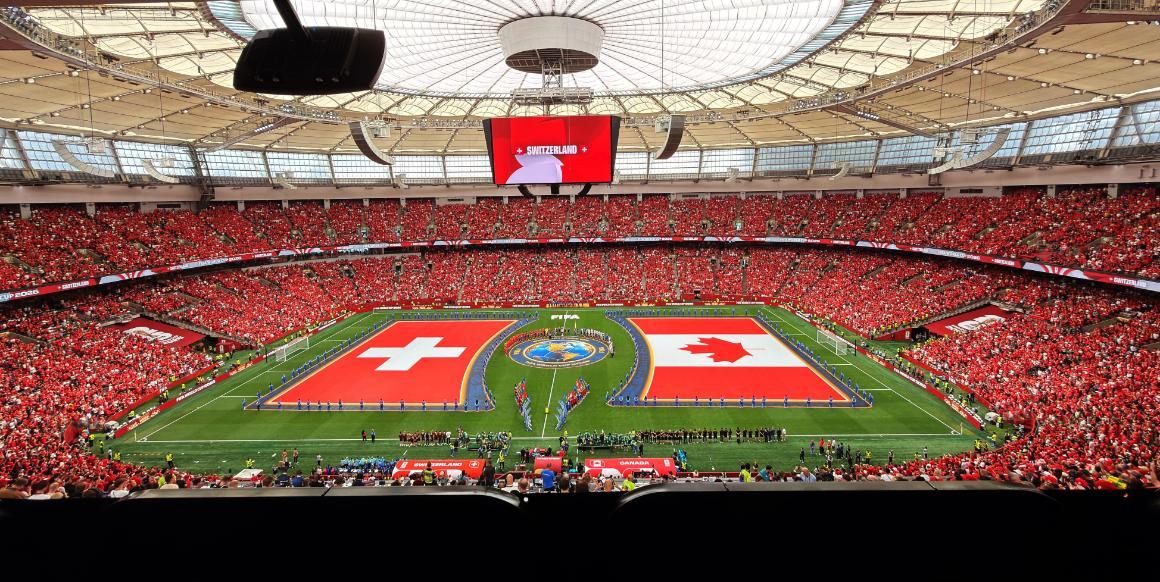 A crowded soccer stadium filled with red-clad fans during a match, featuring the flags of Switzerland and Canada on the field, and a large display screen above.