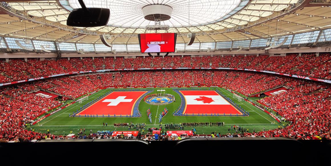 A crowded soccer stadium filled with red-clad fans during a match, featuring the flags of Switzerland and Canada on the field, and a large display screen above.