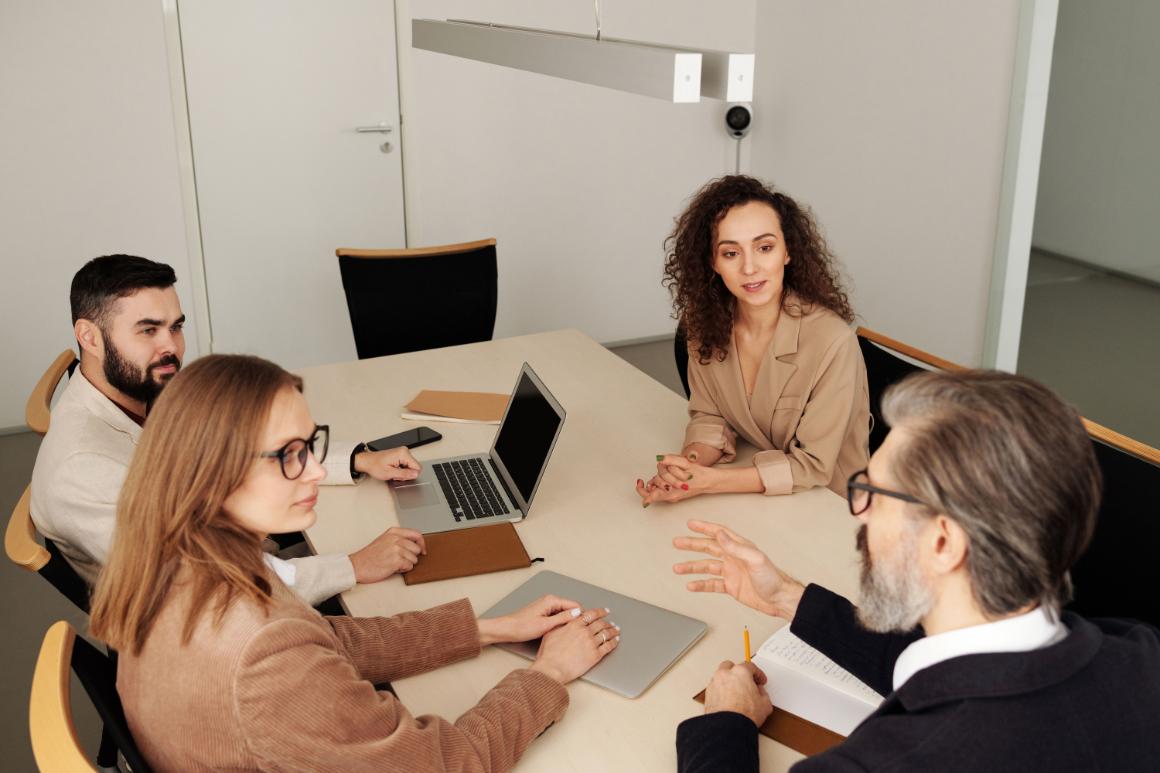 Four adults collaborate around a conference table with laptops, notebooks, and folders in a bright office.
