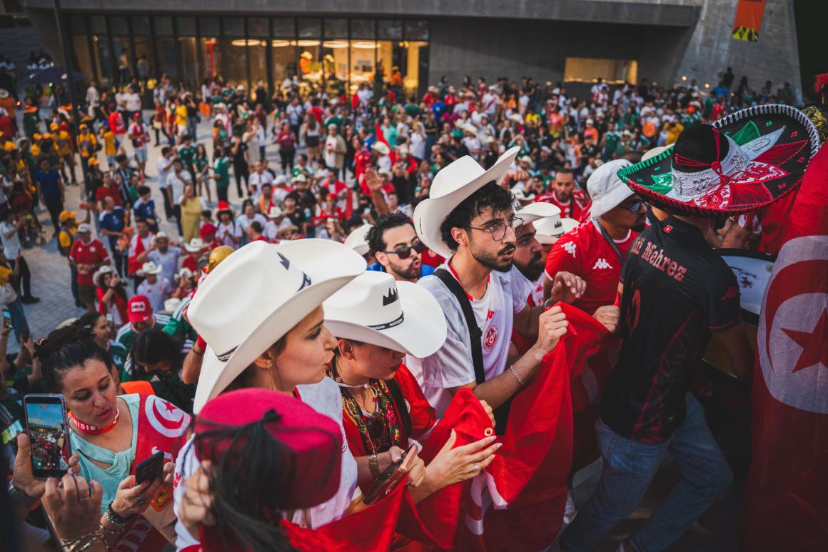A diverse group of people in cowboy hats holds flags and phones at a lively outdoor event with a bustling crowd in the background.