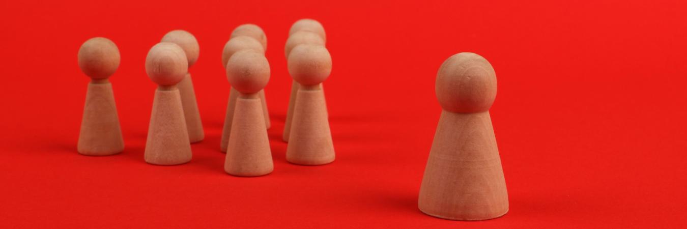 A group of wooden figurines on a red background, with one taller figure standing apart from six smaller figures.